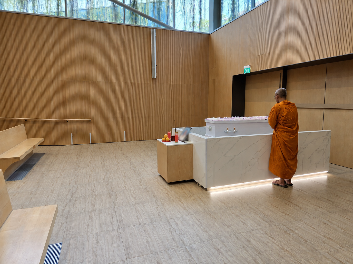 Buddhist monk in orange robes standing beside a white coffin in a peaceful modern funeral hall, with the memorial photo blurred for privacy.