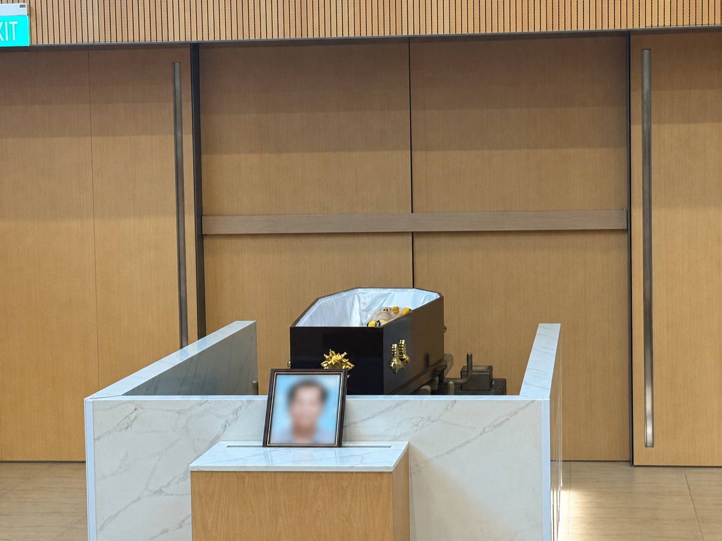 A black coffin positioned inside a modern crematorium hall, with a blurred framed memorial photograph placed in front.