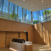 Modern funeral service hall with a black coffin resting on a white marble platform, surrounded by warm wooden walls, tall glass windows, hanging greenery, and natural sunlight.