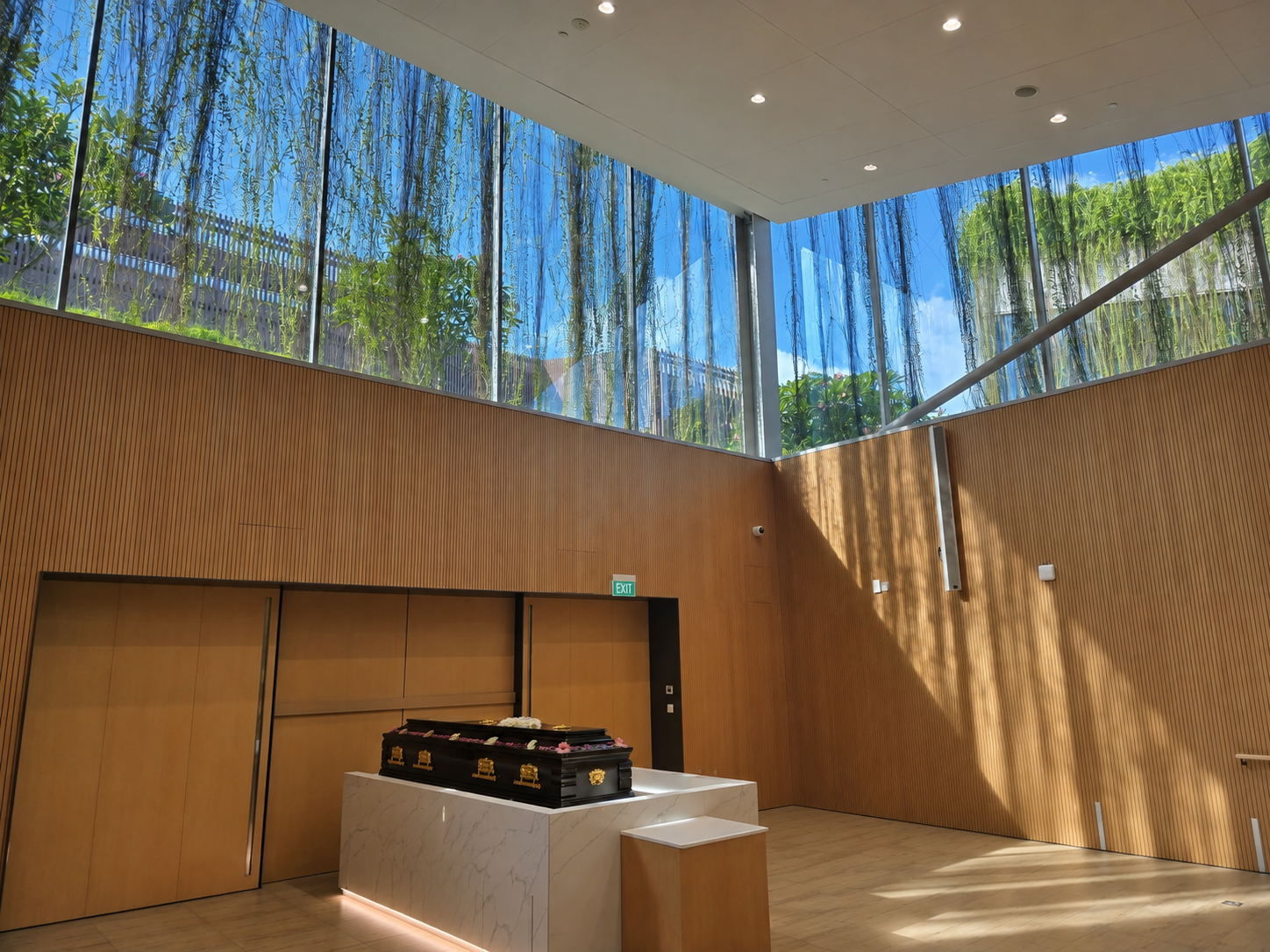 Modern funeral service hall with a black coffin resting on a white marble platform, surrounded by warm wooden walls, tall glass windows, hanging greenery, and natural sunlight.